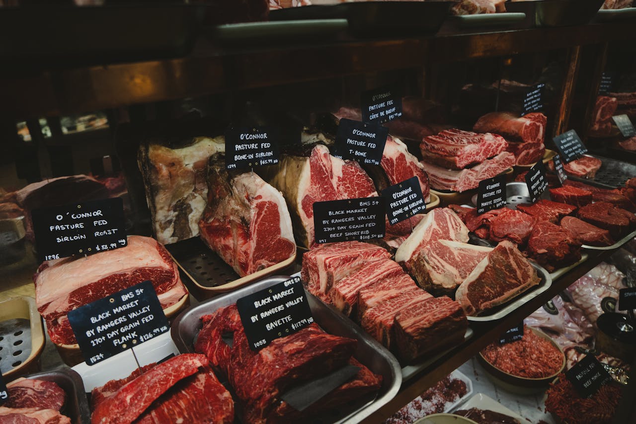 Home A range of raw meat cuts on display in a butcher shop, featuring labels for each type.